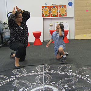 A woman is pretending to be a kangaroo, holding her hands as ears above her head. A young Aboriginal girl watches and mimics.