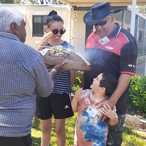 A young Aboriginal boy, a woman and an Aboriginal man are standing together. An elder is covering them in smoke.