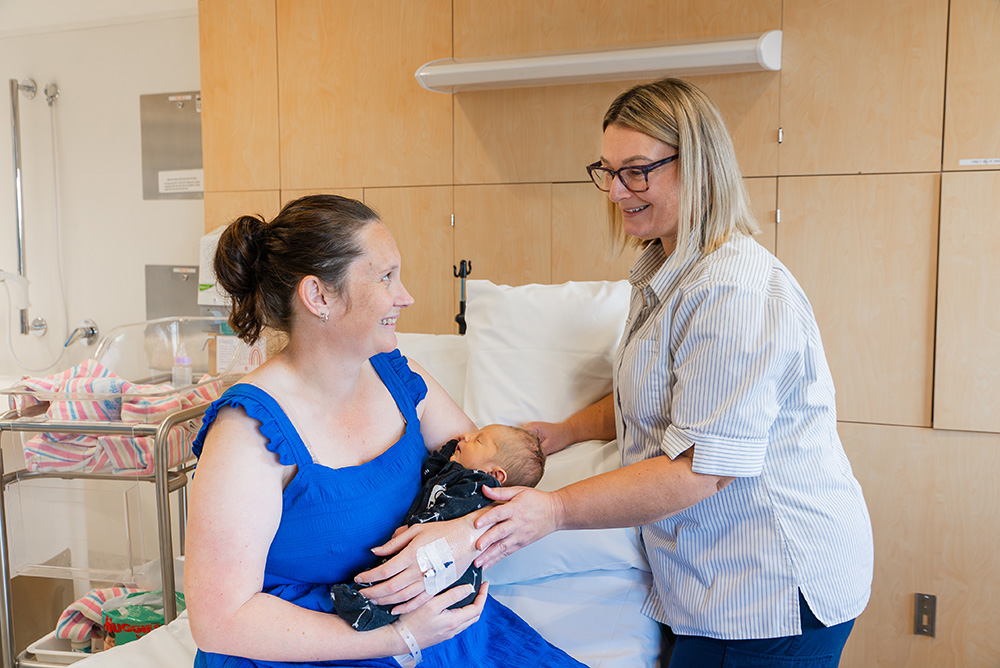 Mother sitting on hospital bed while holding young baby and smiling at midwife